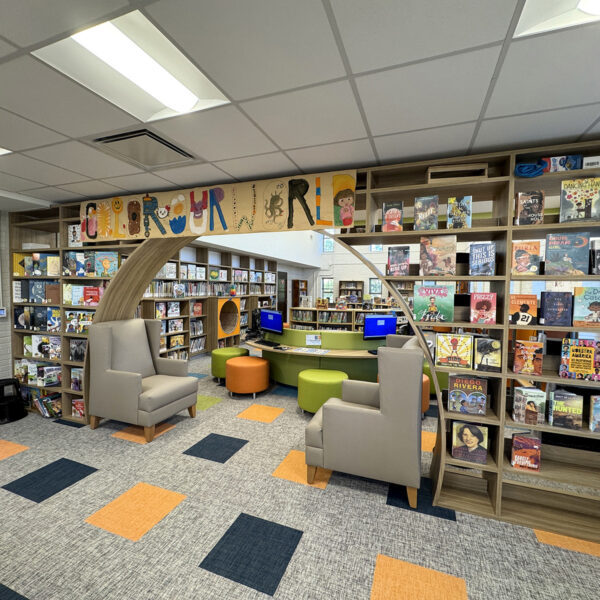 Entrance to the children's area. A wall of shelves with book covers facing forward makes way for a tall semicircular entrance. A banner hanging overhead reads Color Our World.