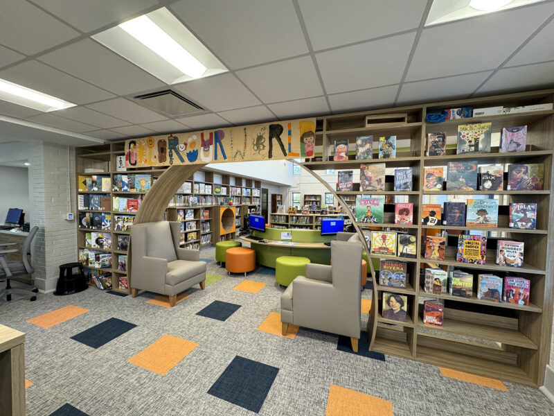 Entrance to the children's area. A wall of shelves with book covers facing forward makes way for a tall semicircular entrance. A banner hanging overhead reads Color Our World.