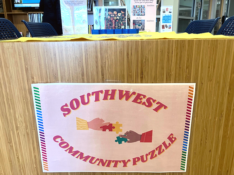 southwest-community-puzzle A table with a sign on it saying Southwest Community Puzzle. The sign shows one hand with dark skin and one hand with light skin, about to connect the puzzle pieces they each hold. On top of the table is a mat with puzzle pieces on it.