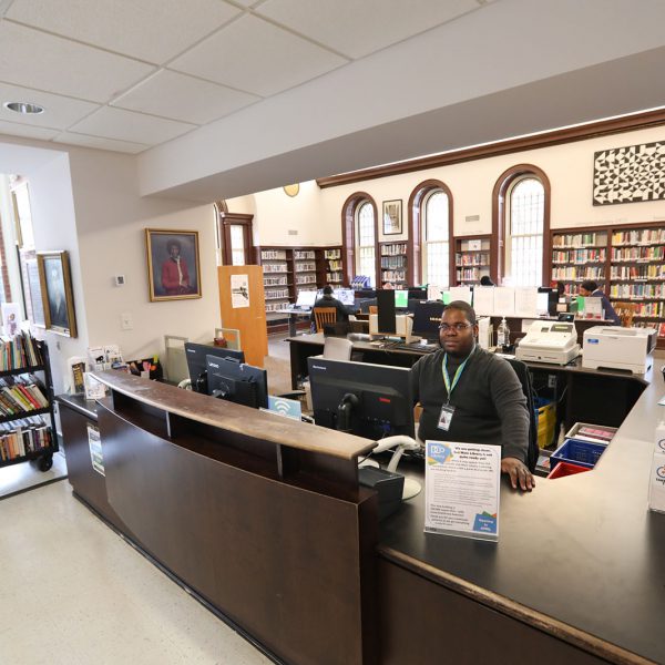 A library staff member sitting at the checkout desk, with the main reading room in the background