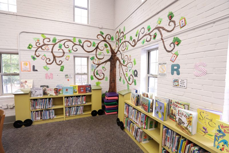 Corner in the children's area with low colorful shelves of books and a tall tree mural branching overhead