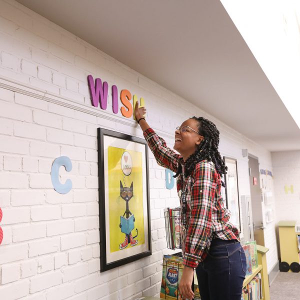 A children's librarian laughs while hanging up foam letters spelling "wish" on the wall