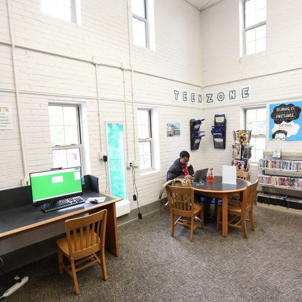 Corner labeled "Teen Zone," with a person sitting at a table using a laptop, a public computer station nearby, and shelves of DVDs