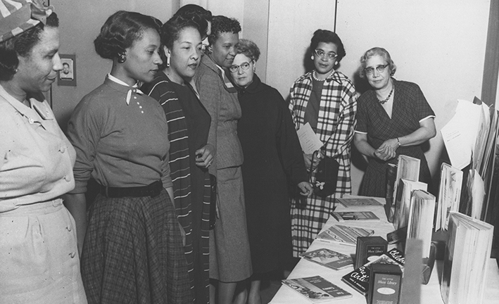 A black and white photo showing a group of Black women in midcentury dresses standing in front of a