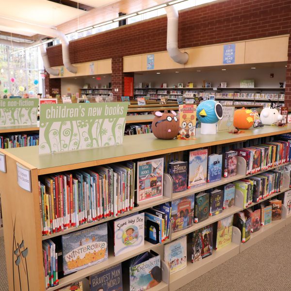 Shelf of children's picture books, with a line of pumpkins decorated to look like different characters along the top and more shelves in the background