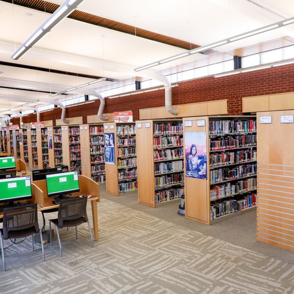A large open room, with computers at desks in front of rows of bookshelves and a wall full of tall windows in the back