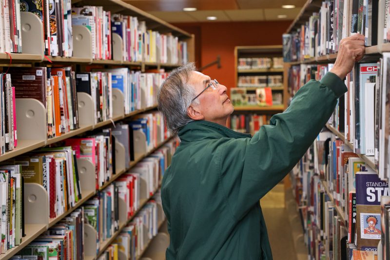 A man stands in the aisle between two bookshelves and reaches up to take down a book