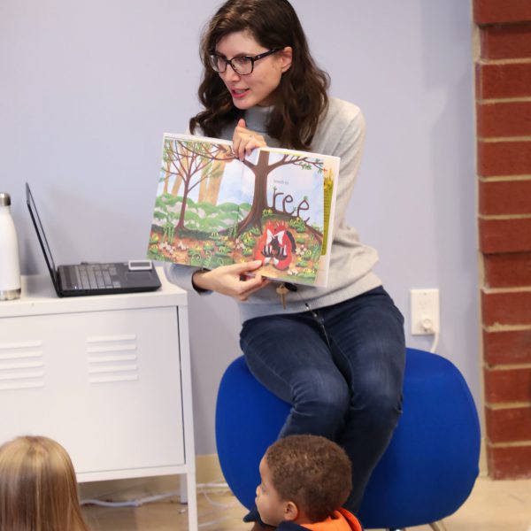 Librarian at the front of the room holding up a picture book as a group of seated children watch