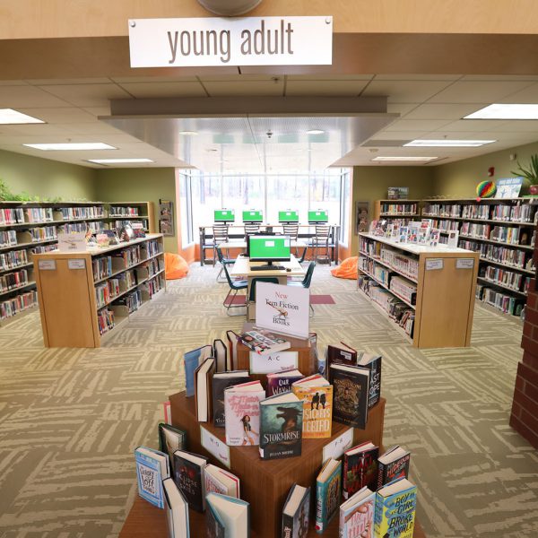 The teen area of East Regional Library, with a display of new teen fiction books at the entrance, walls lined with bookshelves, and computers lined up by a large window at the back wall