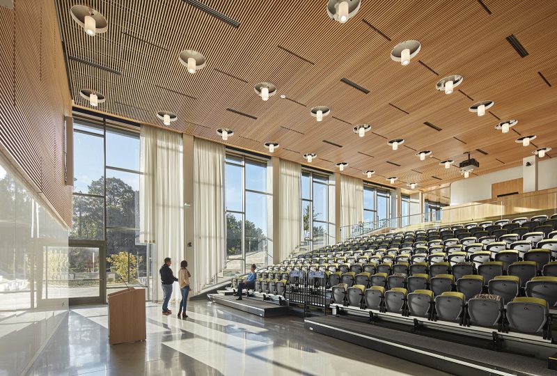 Large auditorium with rows of folded seats, a podium at the front, and a wall of tall windows to the side