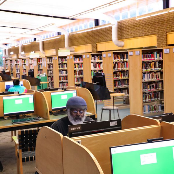 A large open space including a line of desks with computers, many in use, and rows of bookshelves across an aisle