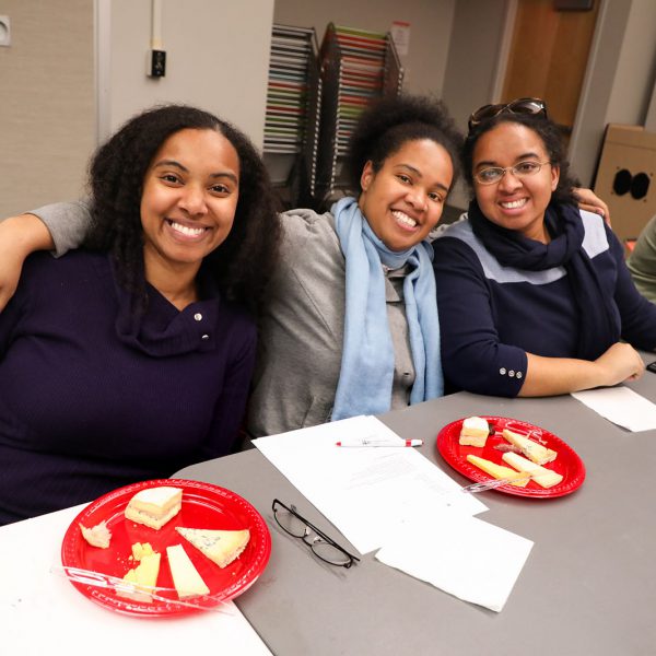 A group of young women smile, each sitting in front of a plate of cheese