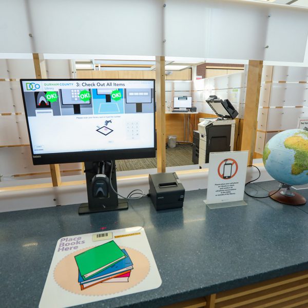 A self-check machine on a counter, with a pad saying "place books here" and instructions showing on a screen. Past the counter are a copier and another 