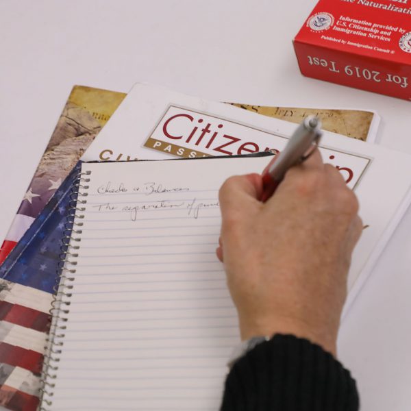 A hand writing in a notebook on top of a book titled "Citizenship: Passing the Test"