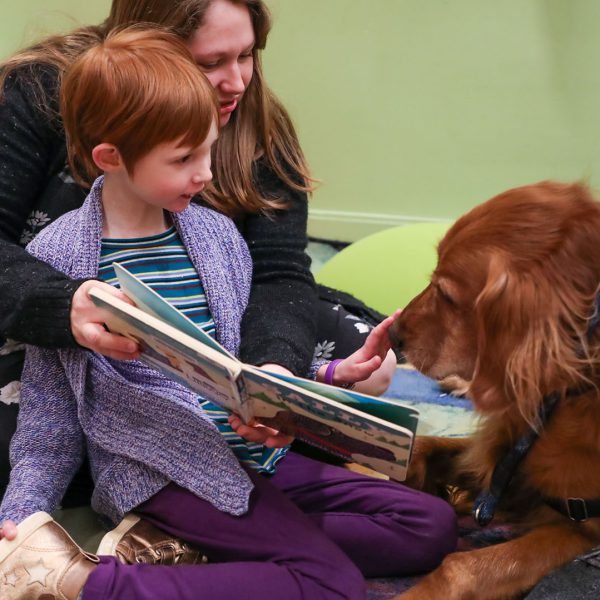 A child pets a visiting therapy dog as her mother holds up a book for them to see