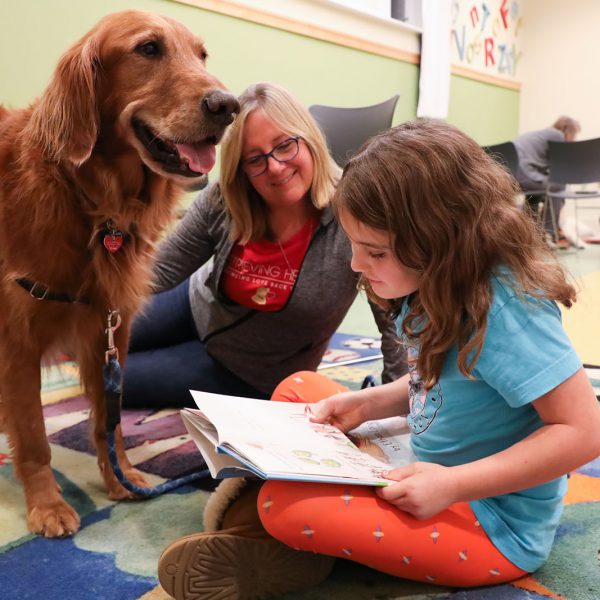A child reads a book to a visiting therapy dog as an adult looks on
