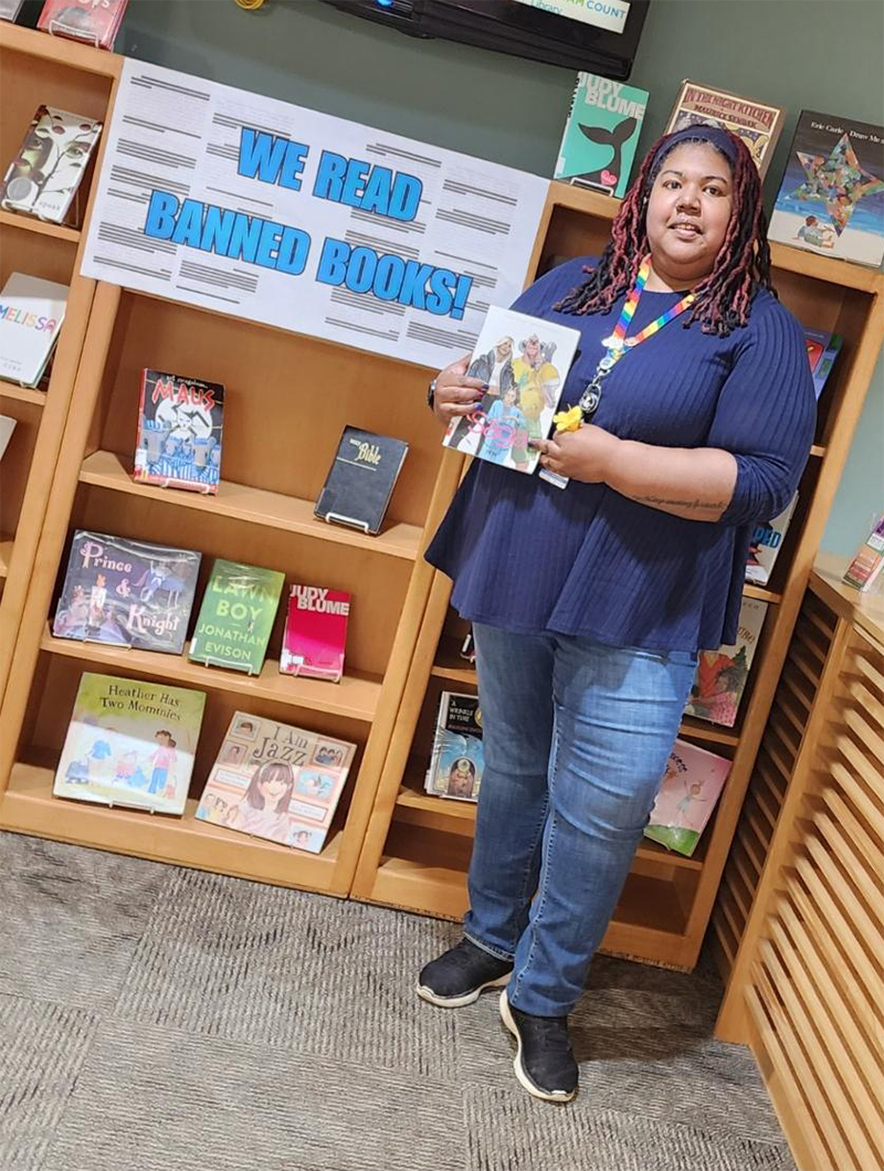 Dominique standing next to a display titled "We Read Banned Books" and holding up the graphic novel "Saga."