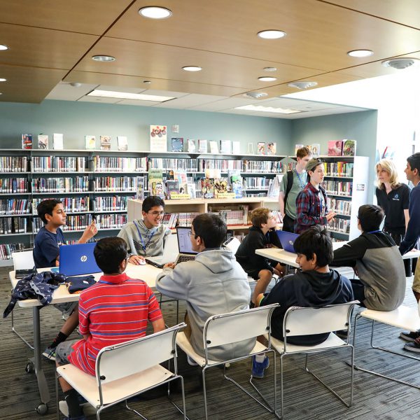 Groups of teens cluster around laptops at a set of tables. Book-filled shelves line the walls behind them