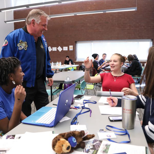 A man in a NASA jacket stands and talks to three teens working at a table