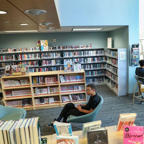 Area with bookshelves, seating, and windows. A teen sits in a chair in the center and looks at his phone