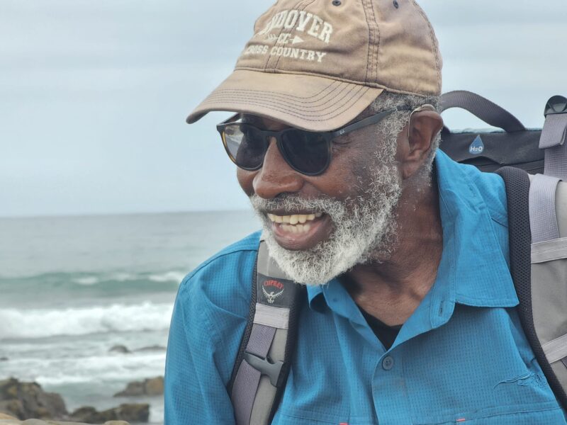 Dr. Francis smiling while walking, wearing a hat, sunglasses, and backpack, with the ocean in the background