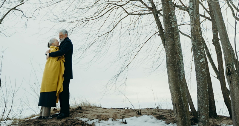 A grey-haired white man and woman embrace in a winter landscape with a grey sky, bare trees, and patches of snow on the ground. Her yellow coat is the only real note of color in the photo.