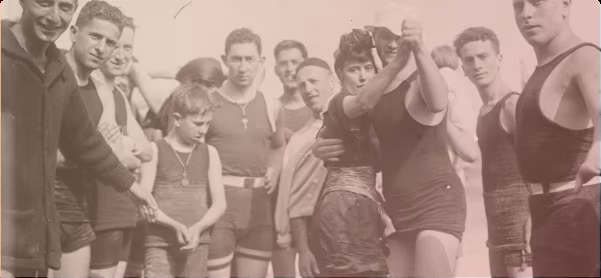 Sepia photo of a group of people of varying ages, with a child and a waltzing couple near the foreground, mostly wearing old-fashioned swimwear but one man wearing a coat.