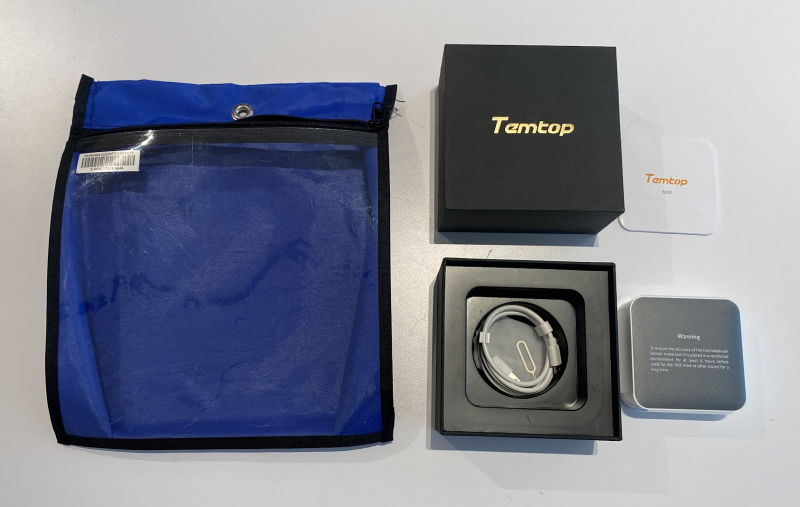 A black box lid saying Temtop, next to the bottom of the box containing a charger cord, next to a small square air quality monitor. The empty blue zippered bag sits next to the kit.