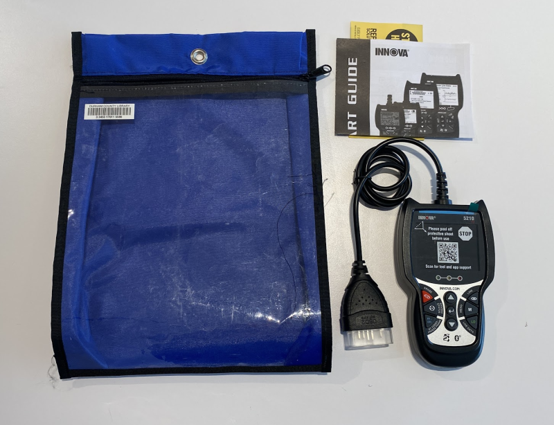 A diagnostic code reader - a black gadget with buttons on the top and a screen covered with a protective sheet including a QR code. The empty blue zippered bag sits next to the kit.