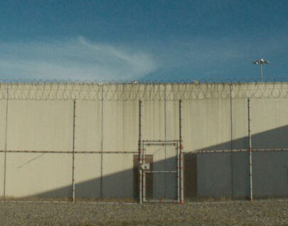 A prison fence with barbed wire on top and a small locked gate, with the grey concrete prison wall behind it and a blue sky overhead.