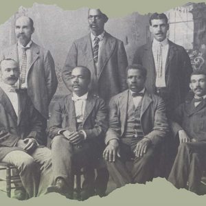 A black-and-white photograph, with ragged edges at the top and bottom, of the founders of the Mechanics and Farmers Bank. They are seven Black men in turn-of-the-century suits and ties, all with mustaches. Four of them sit in the front and the other three stand in a row behind them. Glimpses of the room behind them show patterned wallpaper, a window with what looks like a column on the wall next to it, and flowers.