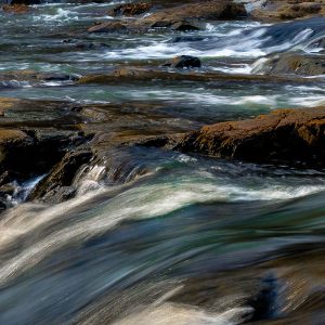 A fairly close-up view of the Eno River flowing over and around brown rocks, with the water looking like a watercolor painting in greens and blues.