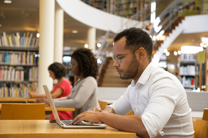 Three adults, each sitting at a separate table working on a laptop in what looks like a library setting. Most clearly visible in the foreground is a light-skinned Black man with a beard and glasses, wearing a white button-up shirt and typing on a silver laptop.