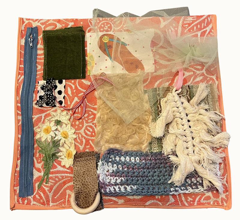 A small blanket with lots of different tactile elements sewn on including a zipper, patch, ring, netting, and more.
