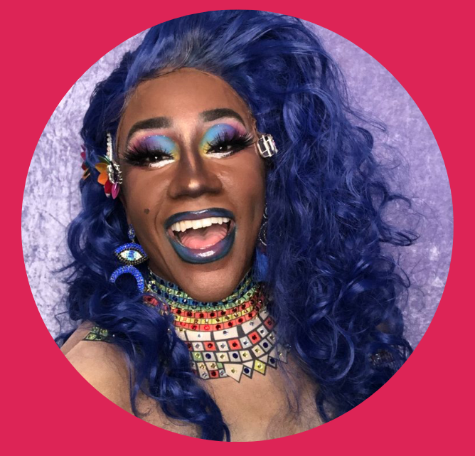 Stormie Daie, a Black drag performer, giving a big smile. She's wearing long purple hair, rainbow eye makeup, and purple lipstick.