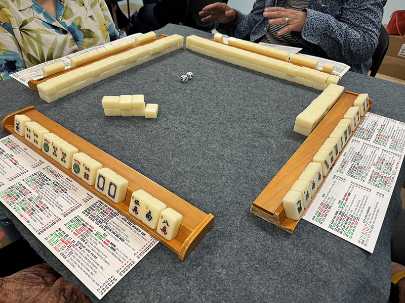 A closeup of a mahjong game, with tiles and cards in the foreground and the backs of the other players' racks visible across the table.