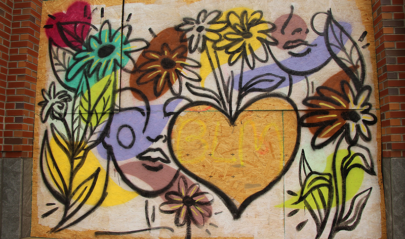 A mural painted on plywood covering a storefront, with large, colorful flowers, outlined faces, and a large central heart unpainted other than the letters BLM in yellow on the plywood.