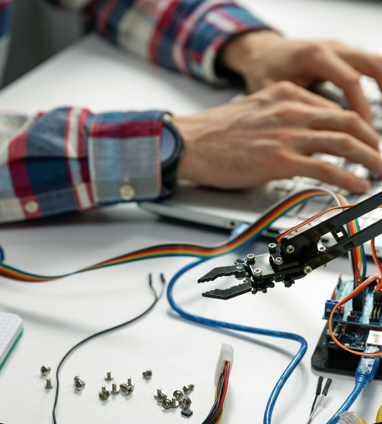 A small hydraulic arm on a white table with screws and cords nearby. In the background a pair of hands can be seen typing on a keyboard.