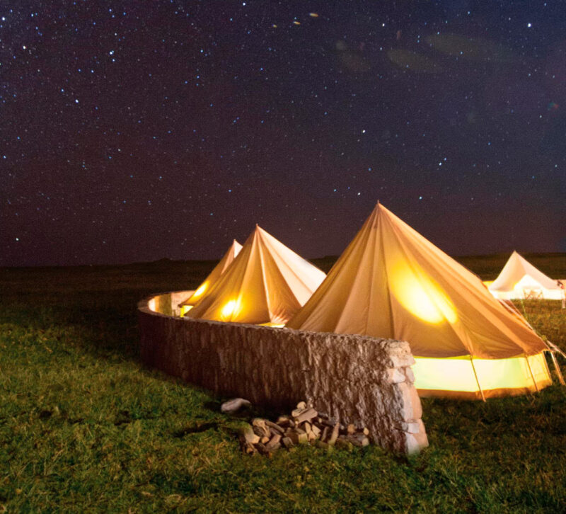 A cluster of tents, each lit from the inside, behind a rock wall on a grassy field under a starry night sky.