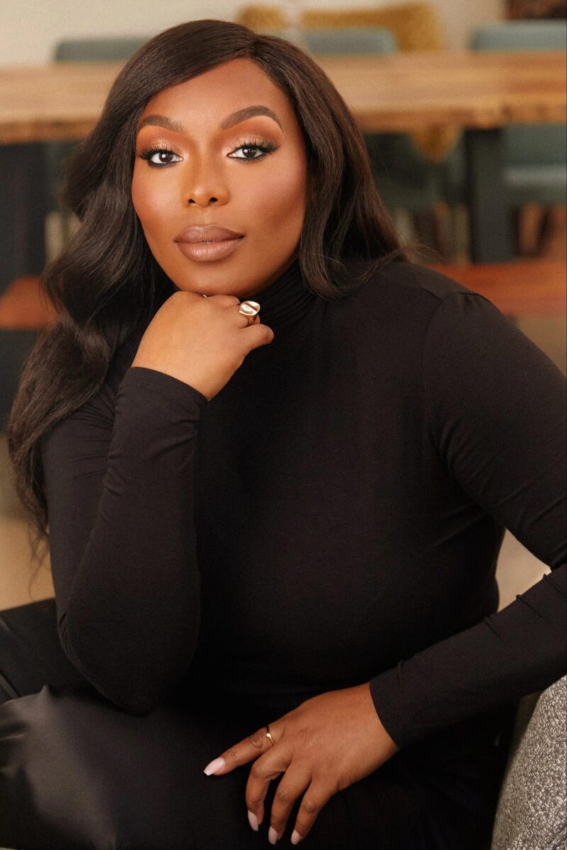 Author Ifeoma Ike, a Black woman with long hair and wearing a black turtleneck, looks at the camera and rests her chin on her hand.