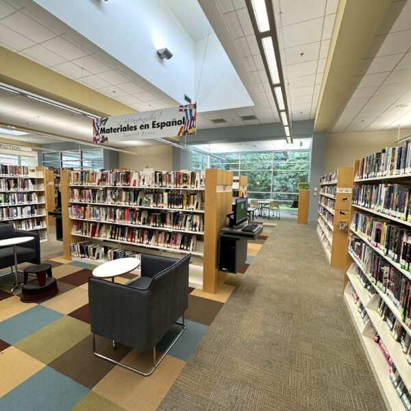 A space with a high ceiling, full of bookshelves with seating in between them and a computer on one of the endcaps. A sign over some of the shelves reads Bienvenidos! Materiales en Español.