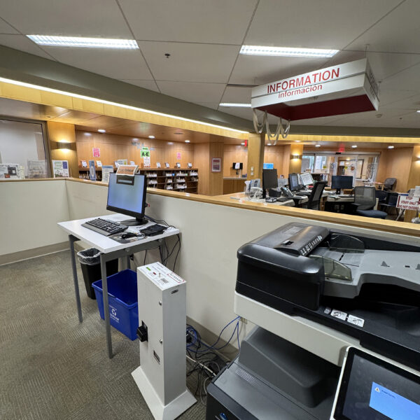 A large copy / printer next to a monitor labeled Print Release Station. In the background, a sign reading Information in English and Spanish hangs over a desk with staff computers and chairs.