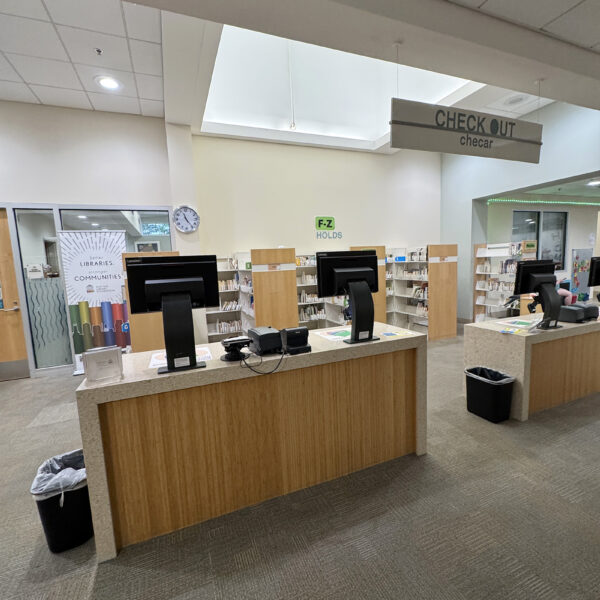 The check-out area, with two tables set up with self-checkout machines, in front of a set of bookshelves full of items on hold.