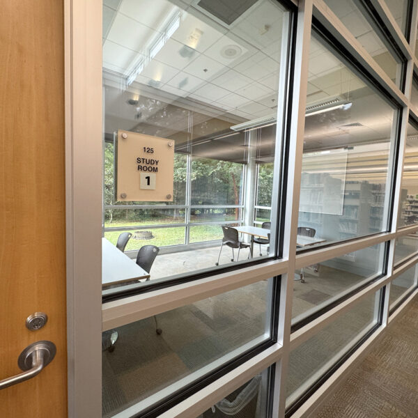 An empty room with two tables, chairs, and a white board, seen through a glass wall with a sign reading Study Room 1.