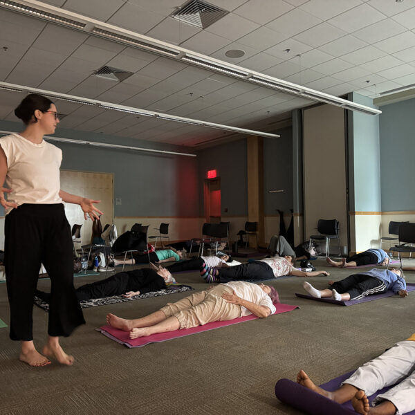 A darkened room full of people of different races in a rest pose on yoga mats. The instructor is walking among them and giving instructions.